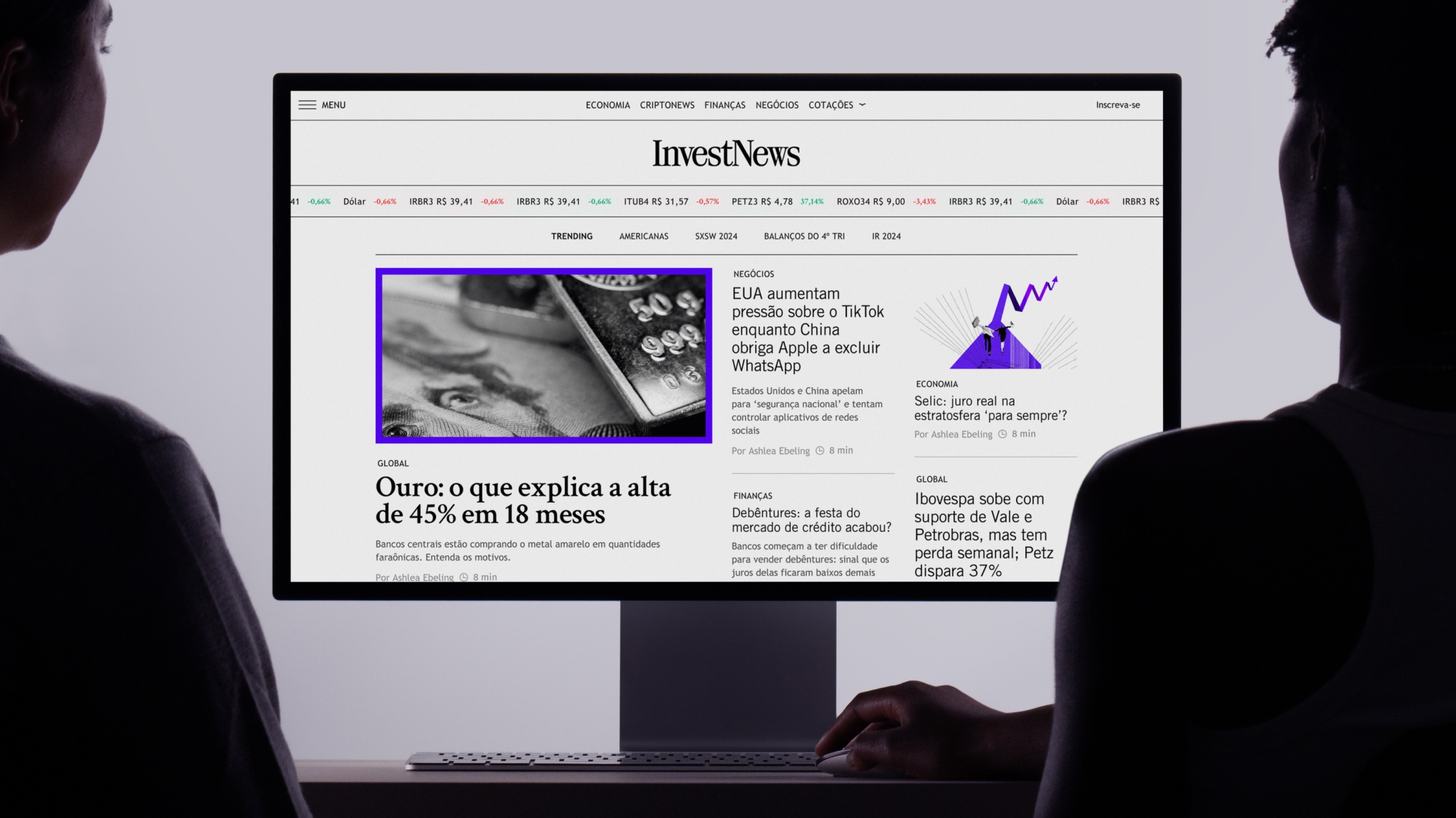 Two people at a desktop monitor showing the InvestNews homepage with its ticker and headline grid