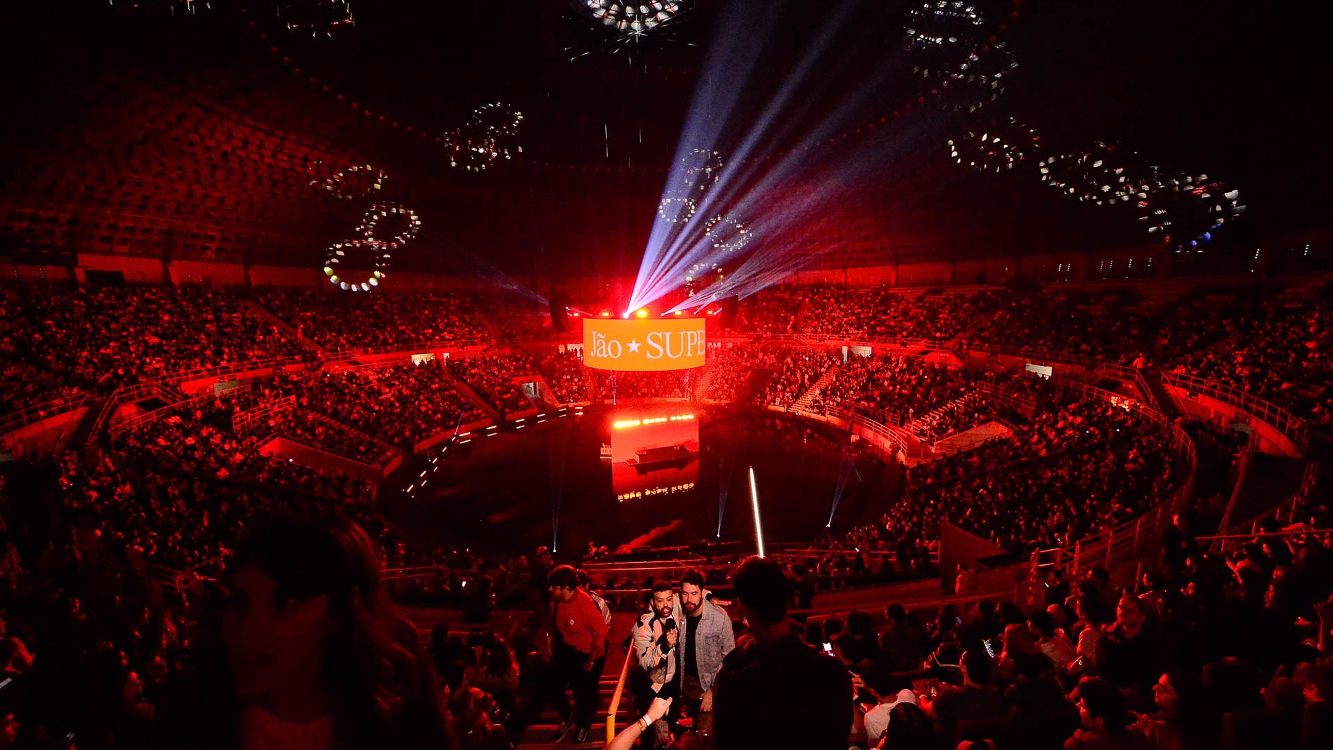 View from the stadium crowd toward the stage, with the "Jão Super" sign lit orange above the floor